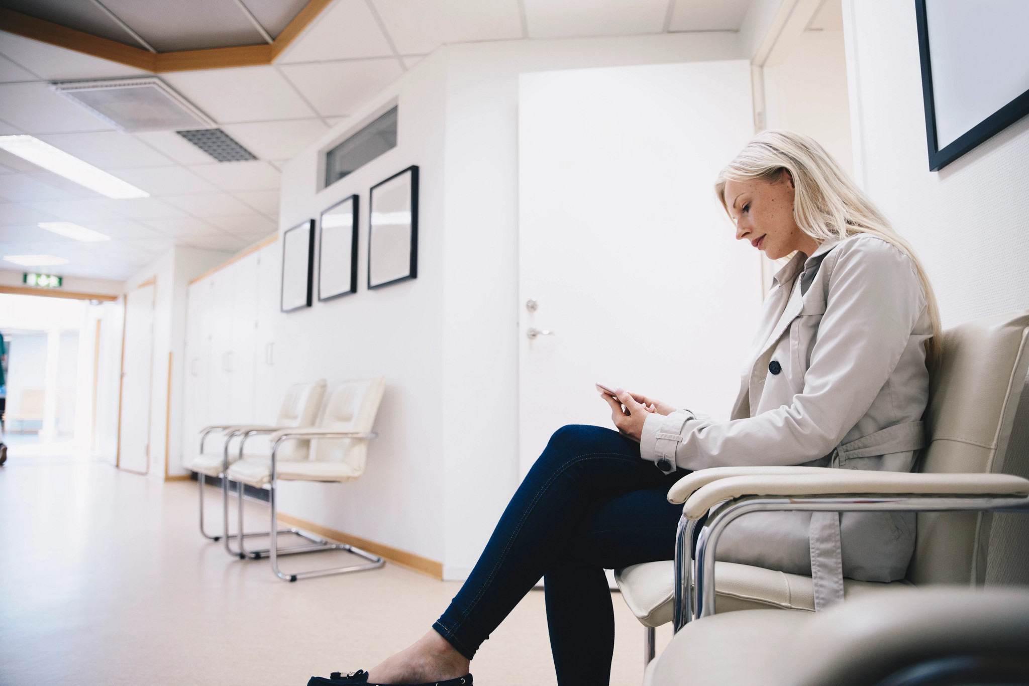 Side view of woman using smart phone while sitting on chair in hospital corridor