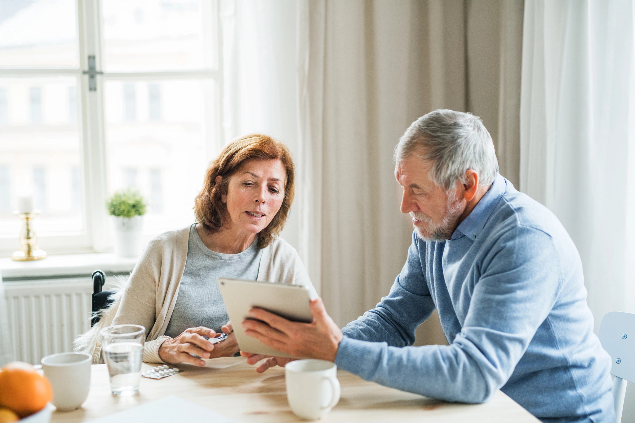 An elderly man and his disabled wife with tablet indoors, explaining how to take medication.