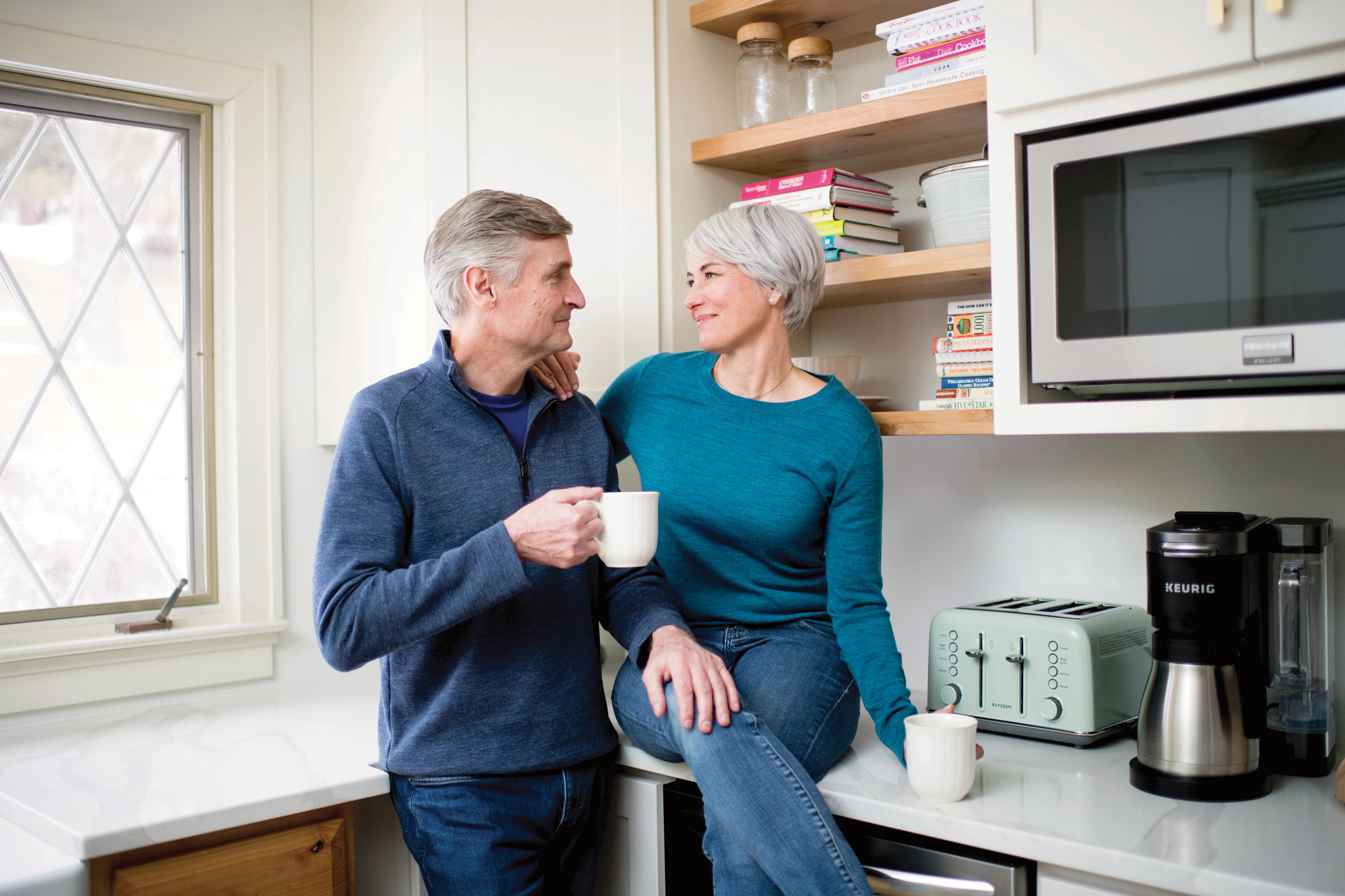 Senior couple smiling and looking at each other while holding coffee in the kitchen.