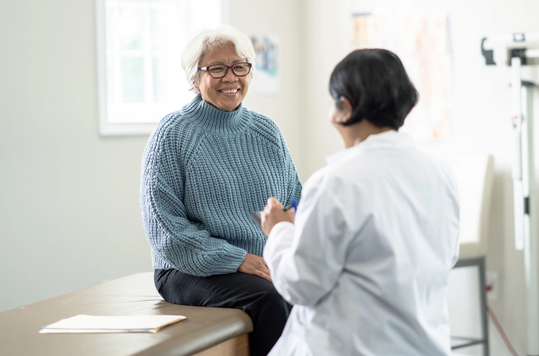 A senior woman sits up on the exam table at a doctors appointment.