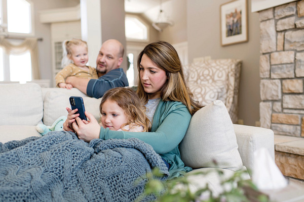 Concerned mother holding sick daughter on a couch with a tablet, while husband and younger daughter look on.