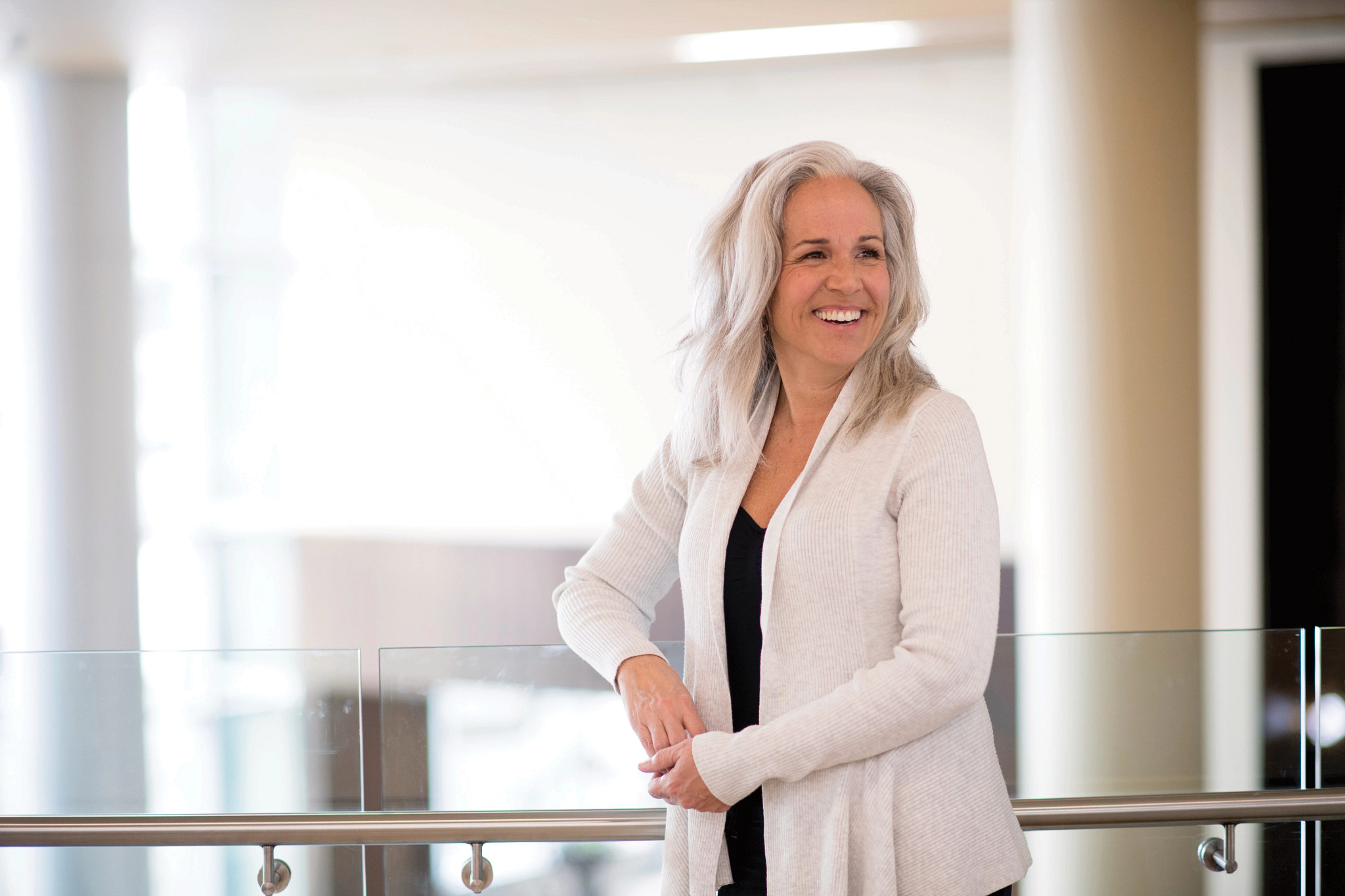Adult woman in a white cardigan leaning against glass smiling.