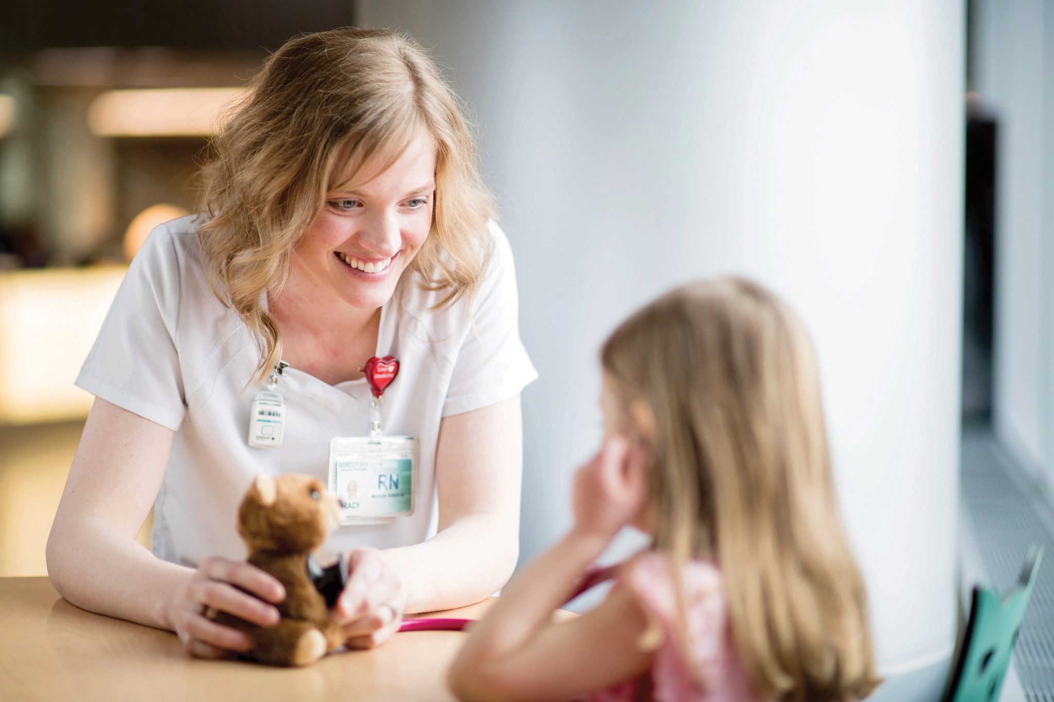 Adult female nurse holding stethoscope to teddy bear with young female patient.