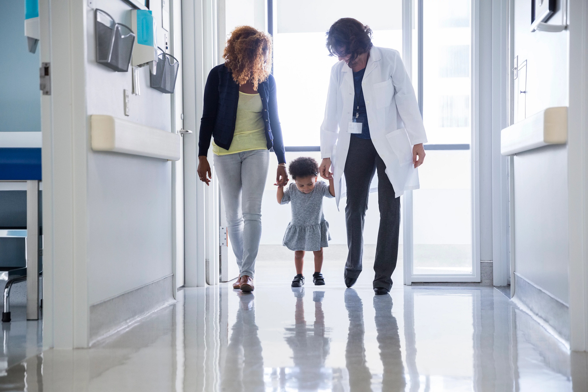 Mother and pediatrician holding hands of toddler while walking in hospital corridor.