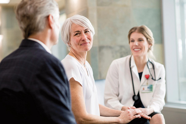 Mature wife looks lovingly at mature husband with young adult female doctor looking on.