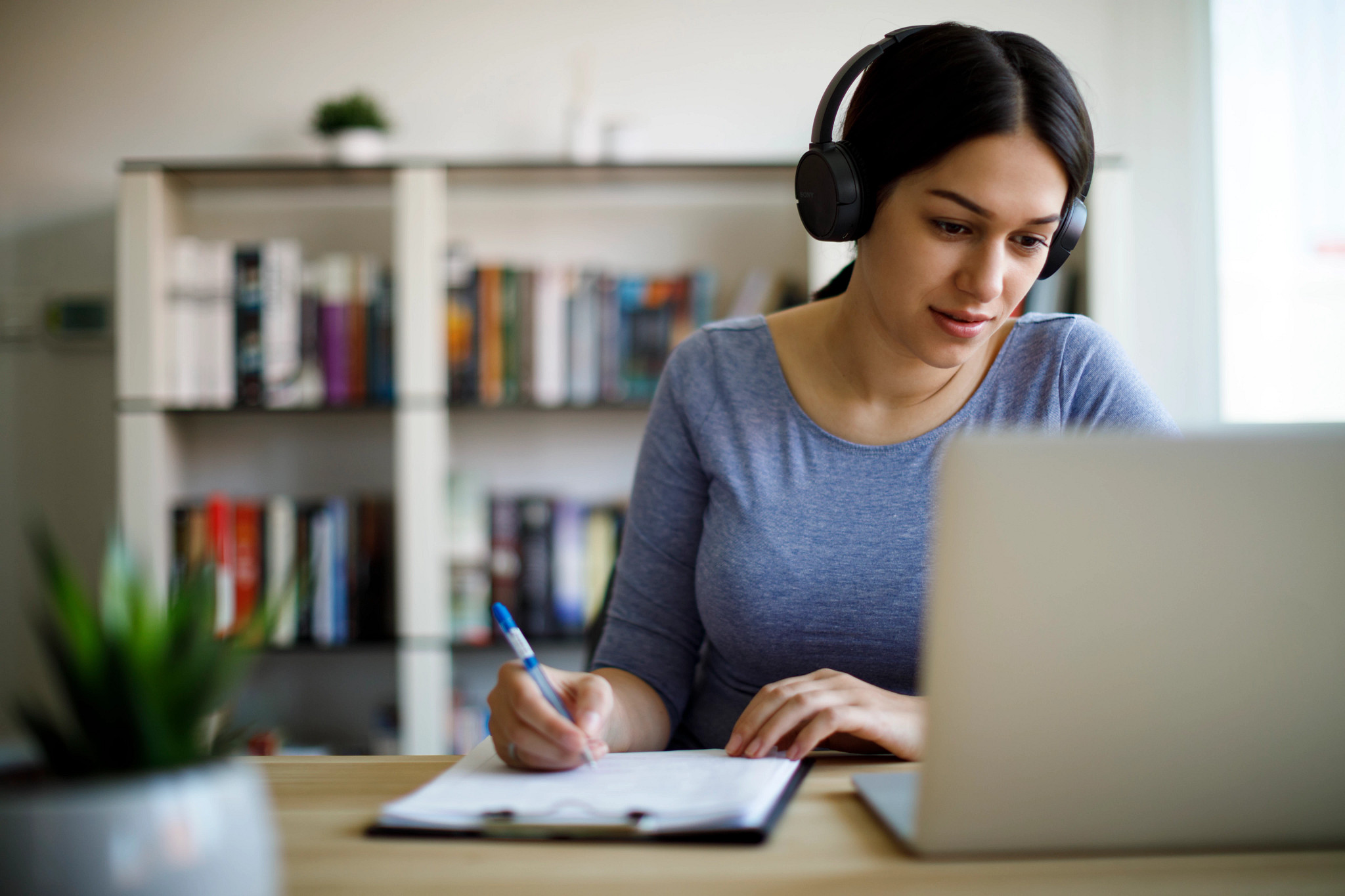 Woman studying at laptop with headphones on and writing on notepad..