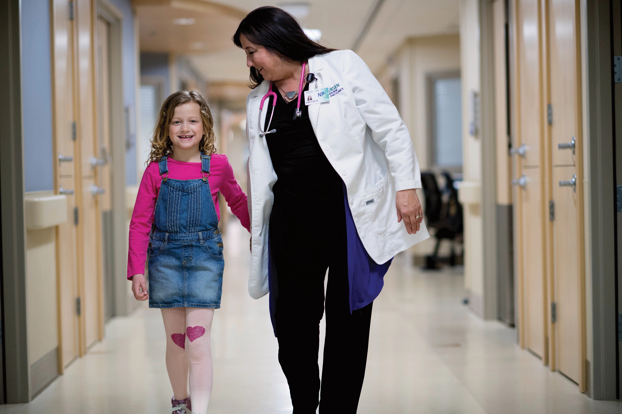 Adult female nurse practitioner walking with young female patient down hospital hallway.
