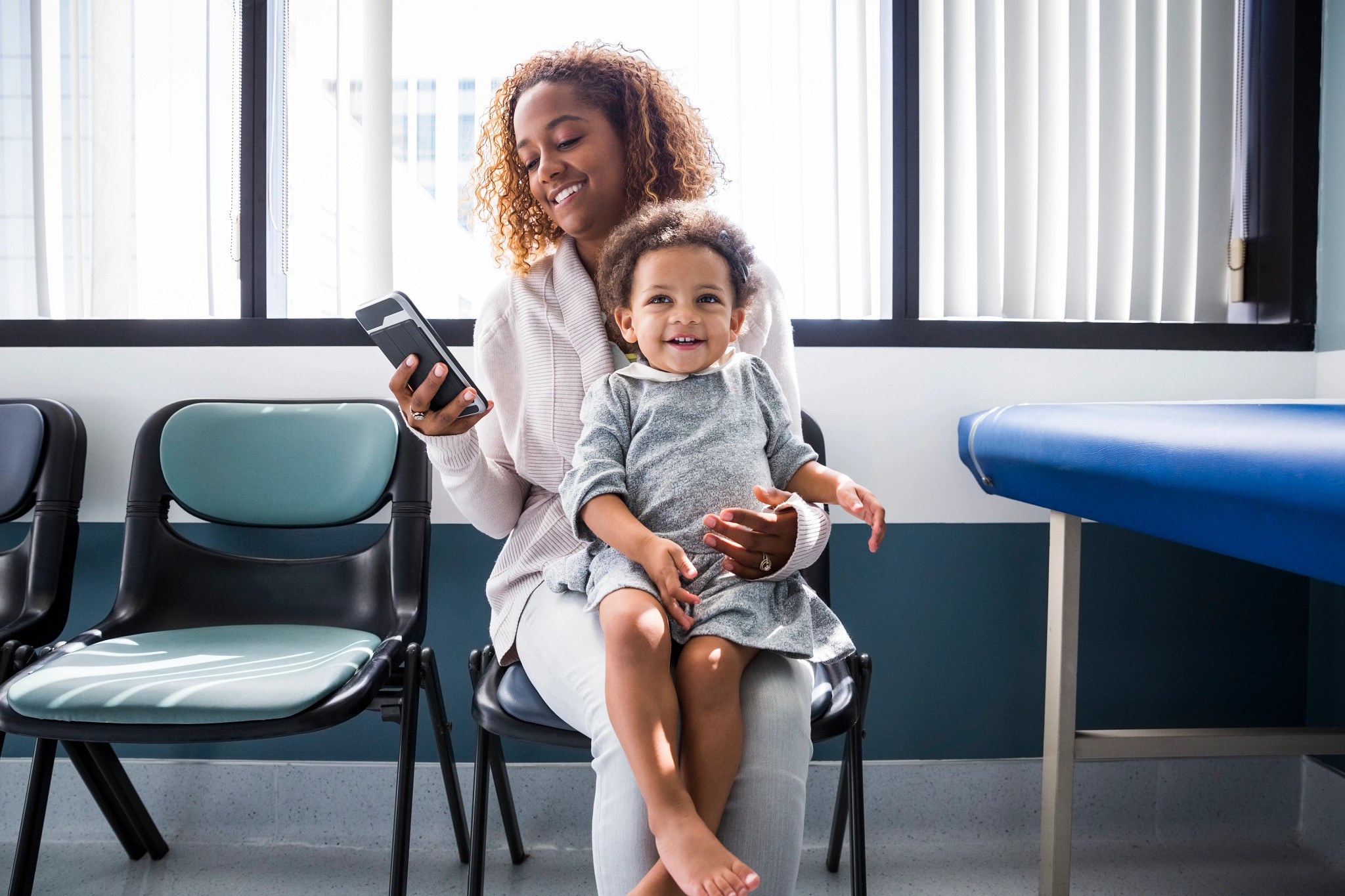 Mother using smart phone while toddler sits on lap at the doctors office.