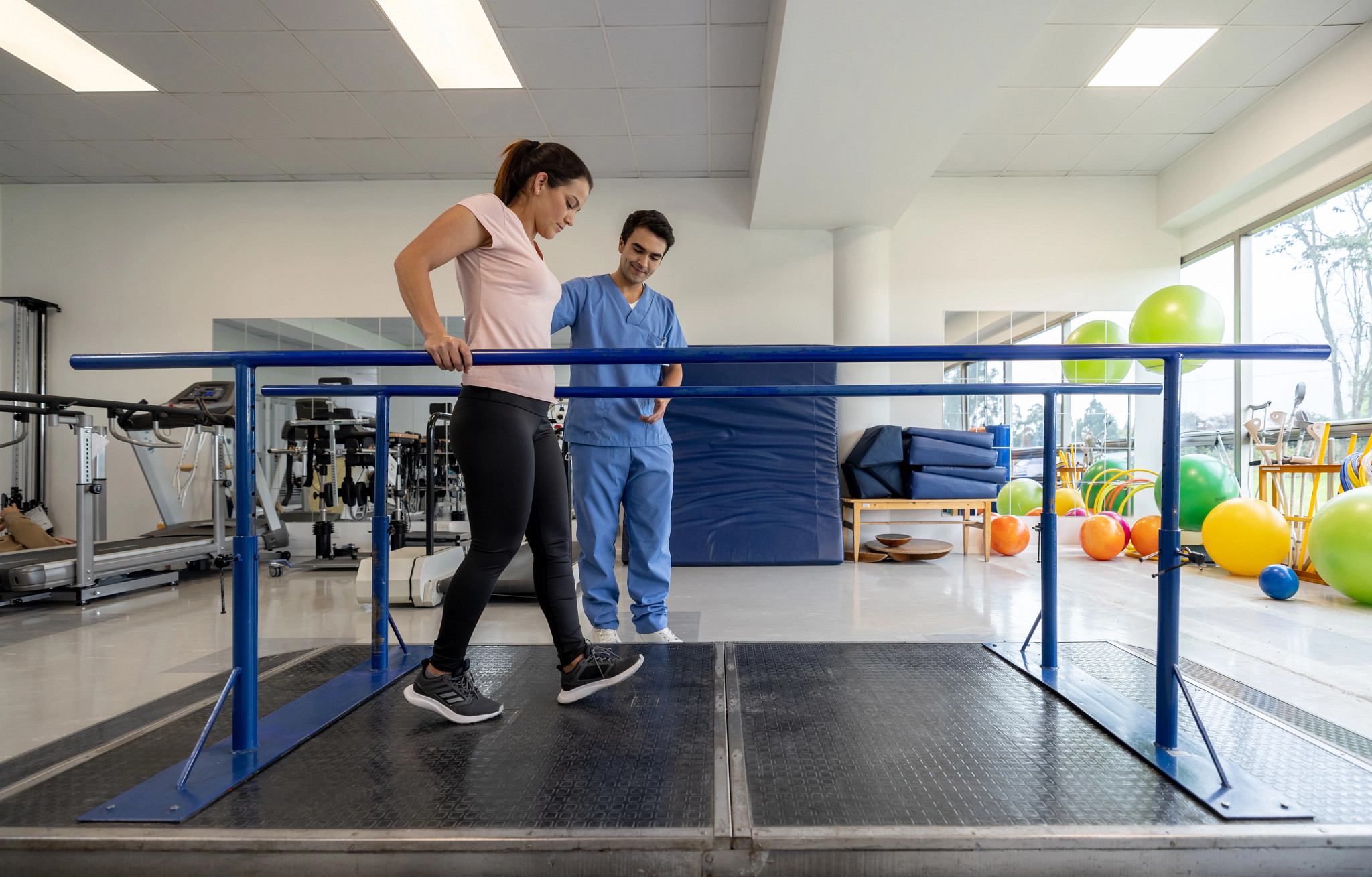 Latin American woman in physical therapy walking on the parallel bars with the assistance of his therapist.