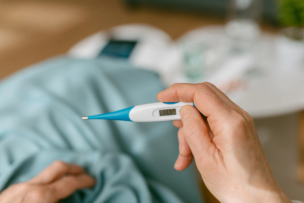 Over-the-shoulder view of an unrecognizable woman looking at the thermometer because she is sick while sitting on the sofa covered with a blanket.
