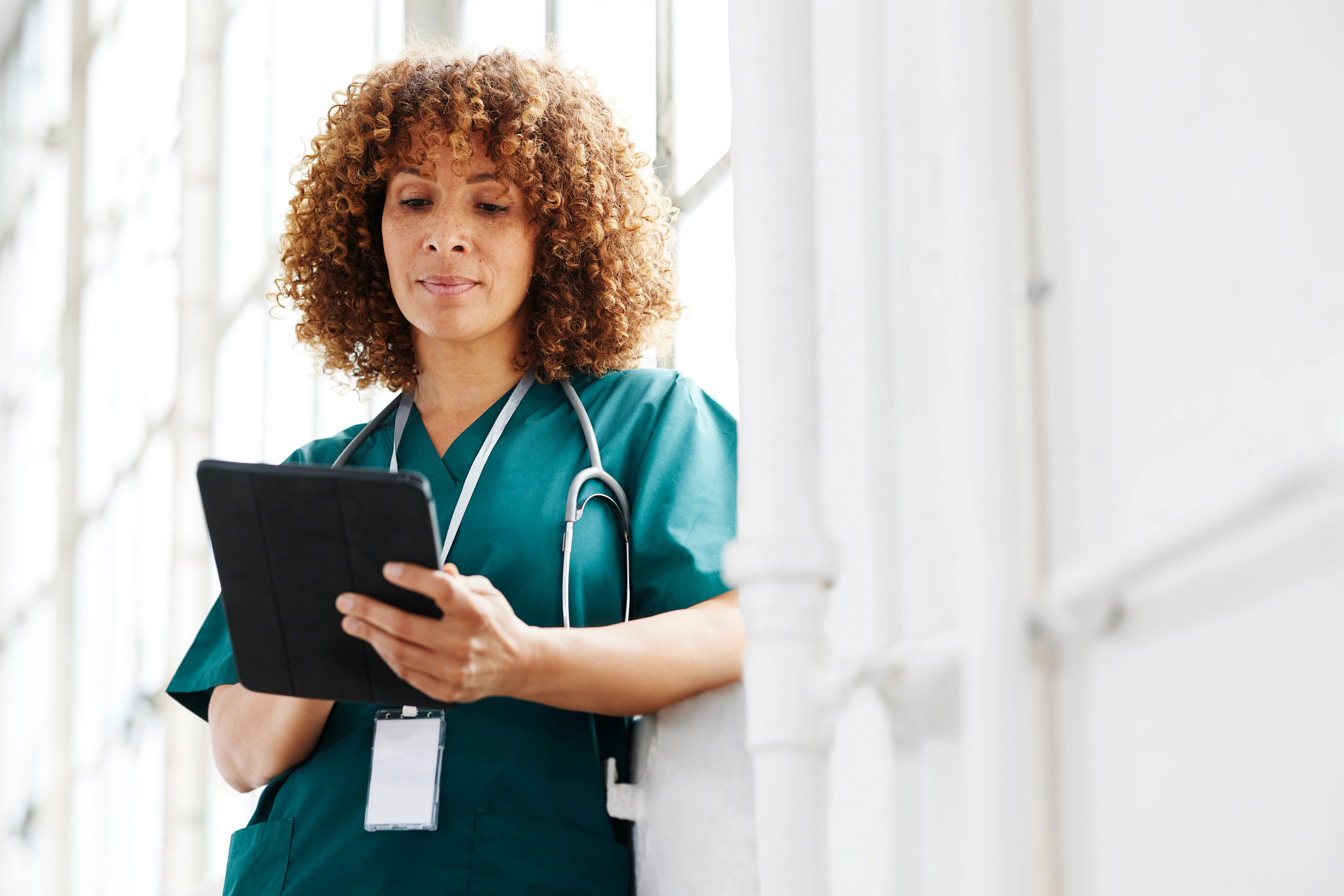 Female healthcare professional looking at digital tablet leaning against window.